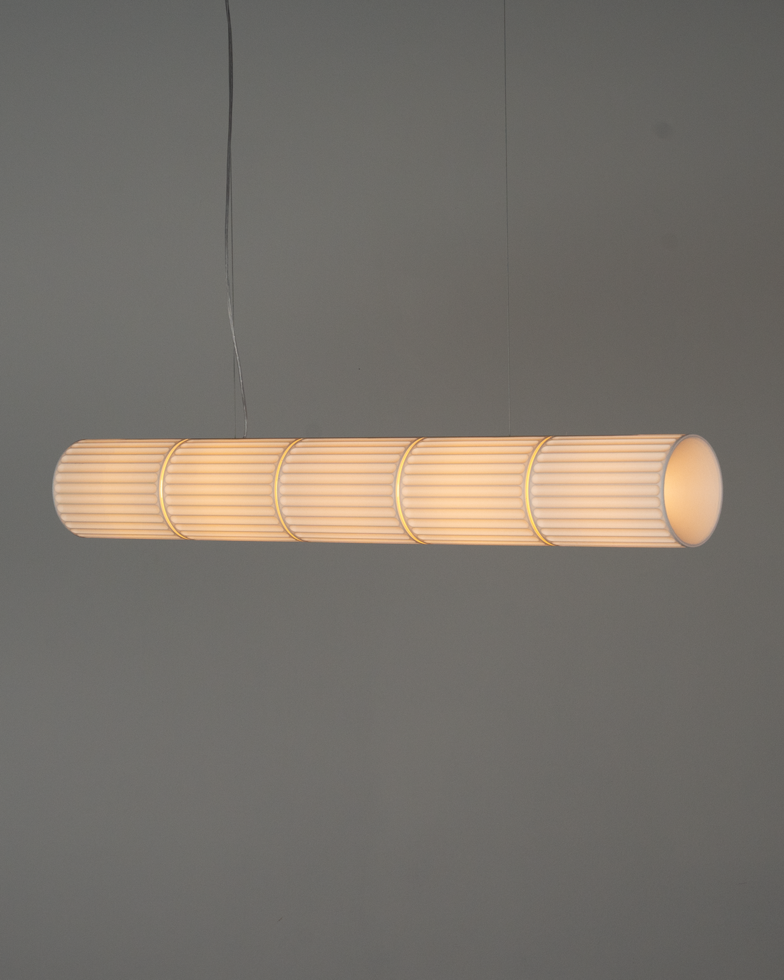Pendente PAR de cinco cúpulas aceso, pendurado por cabos finos diante de uma parede cinza, com luz quente atravessando as cúpulas caneladas.